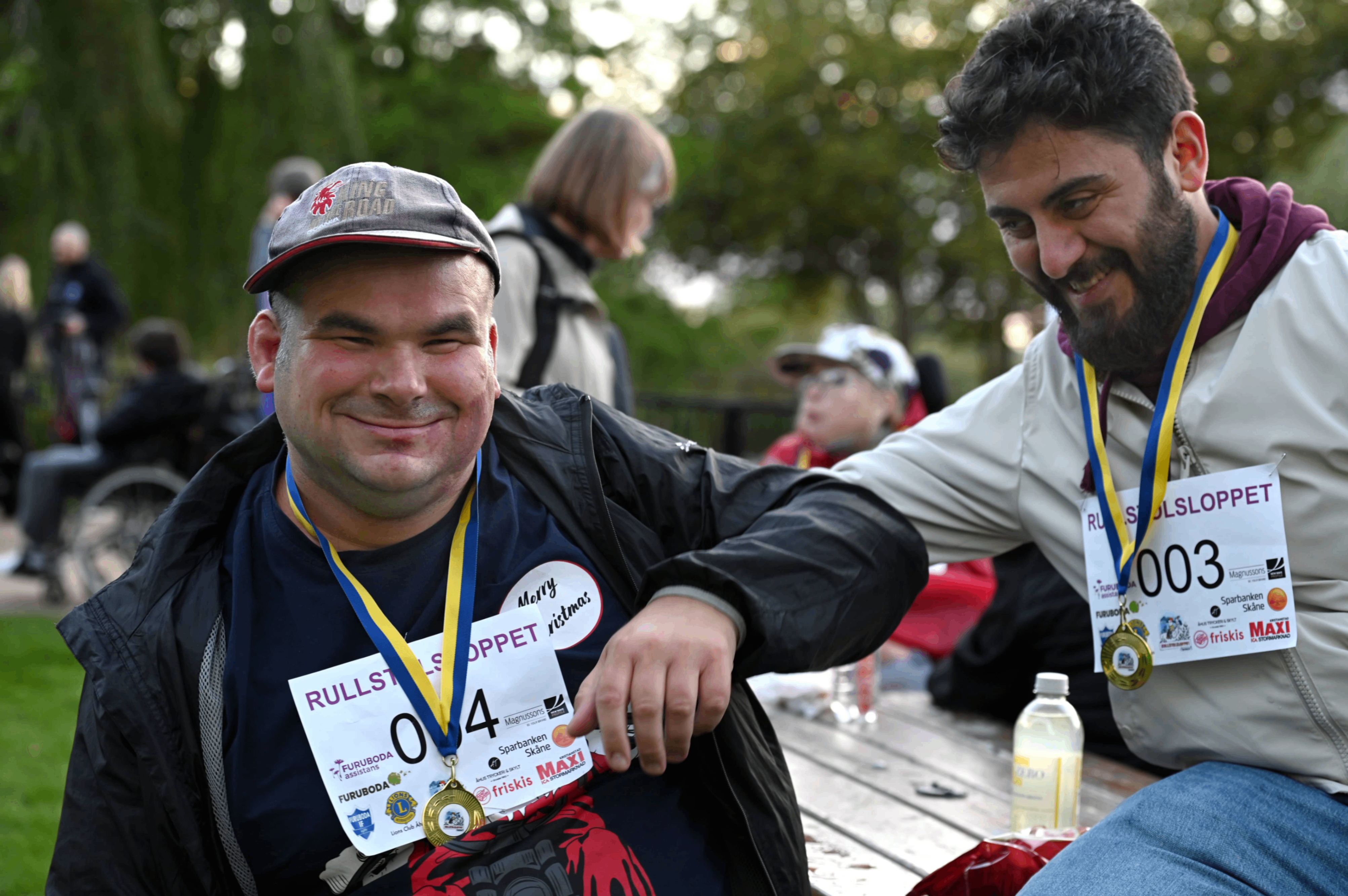 En manlig deltagare med ett stort leende. Har en medalj runt sin hals. Bredvid honom sitter en glad manlig personlig assistent med medalj runt halsen. 