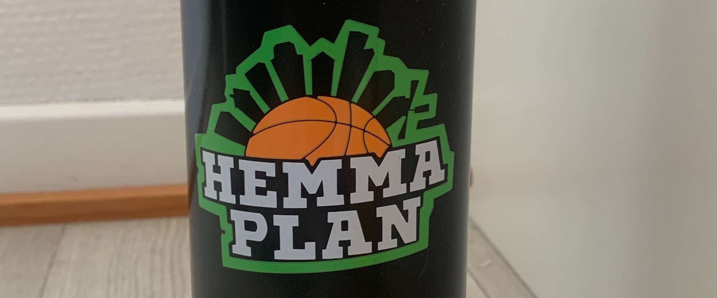 Hemmaplans logga på mörk bakgrund. Loggan består av en halv orange basketboll på en stads siluett där bakgrunden är grön och siluetthusen är svarta. Under baksetbollen står det HEMMAPLAN med vit text