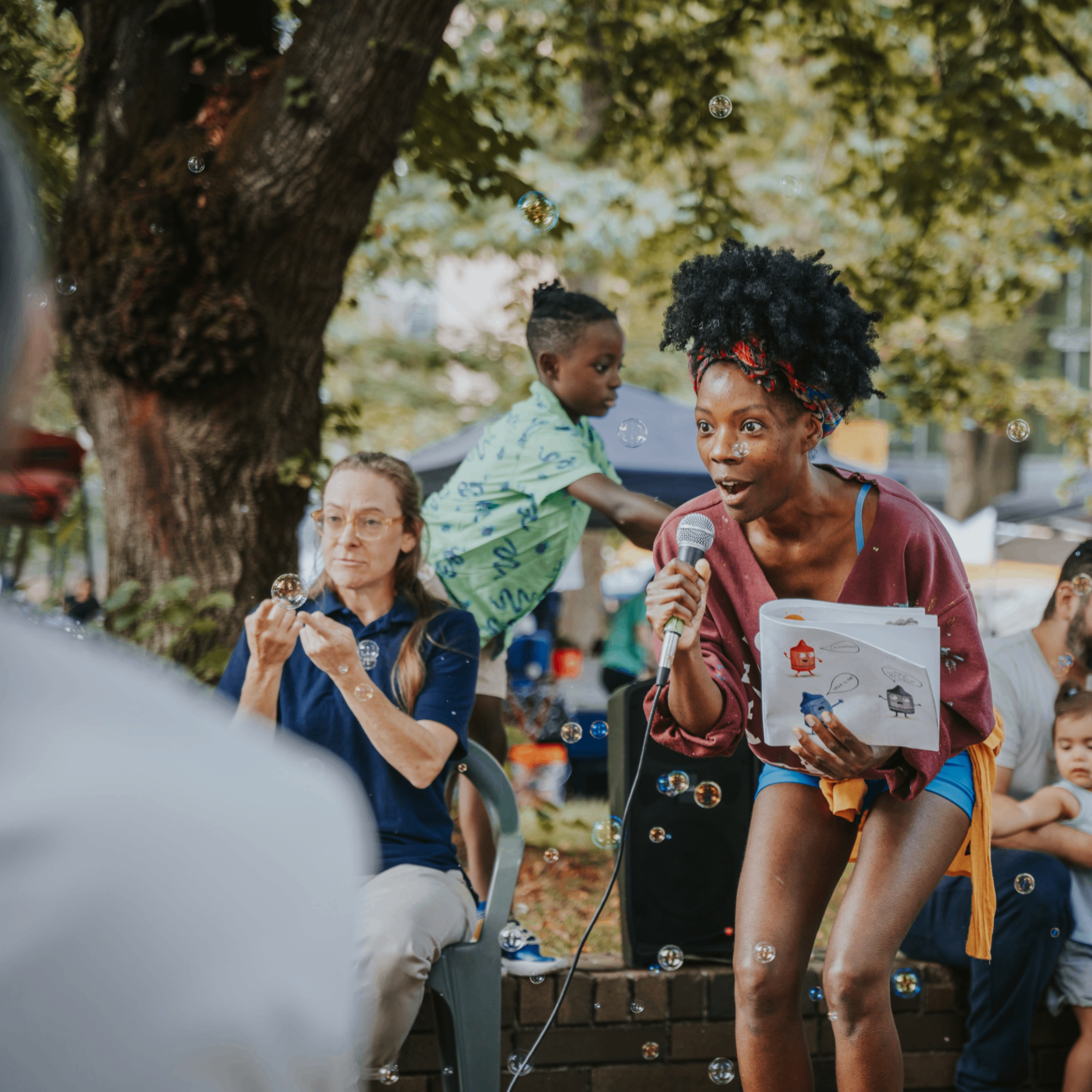 A woman stands with a microphone and a children's illustrated storybook in hand at a farmers market