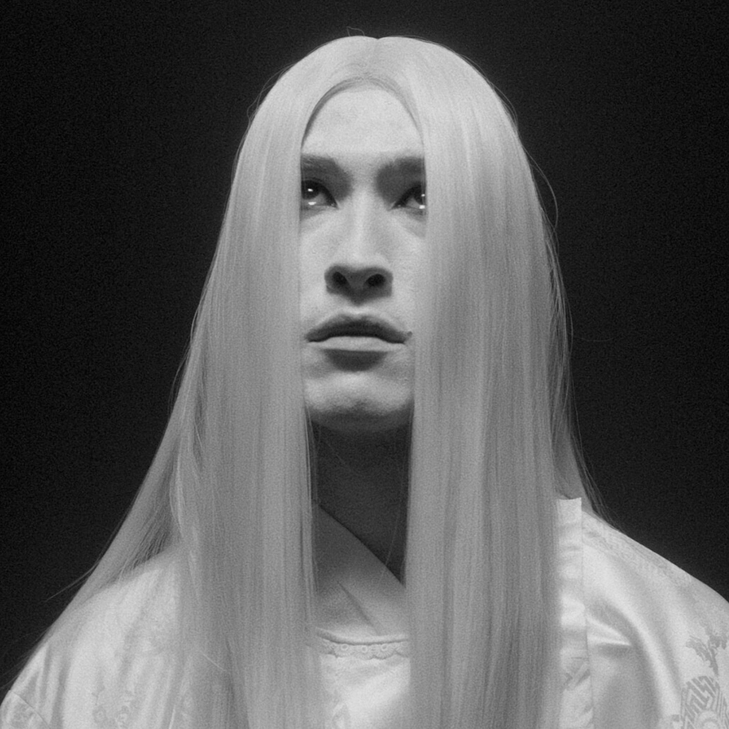 The head and shoulders of a Japanese actor in white clothing and makeup stands against a black backdrop.