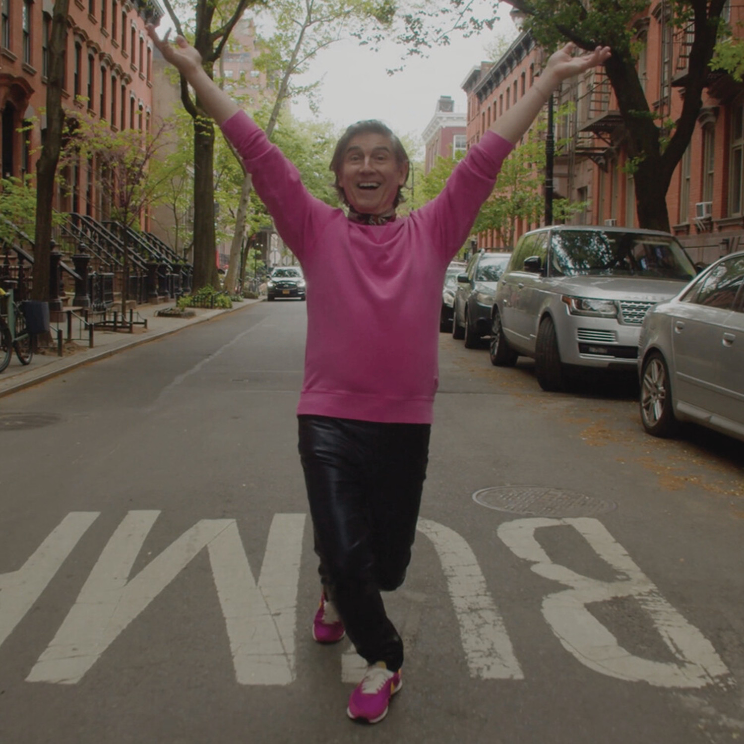 Celeste Lecesne dressed in a pink top stands in a New York street with their arms raised in a V shape.