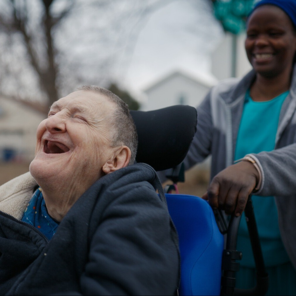Older patient in wheelchair being pushed by caregiver outside. Both laughing.