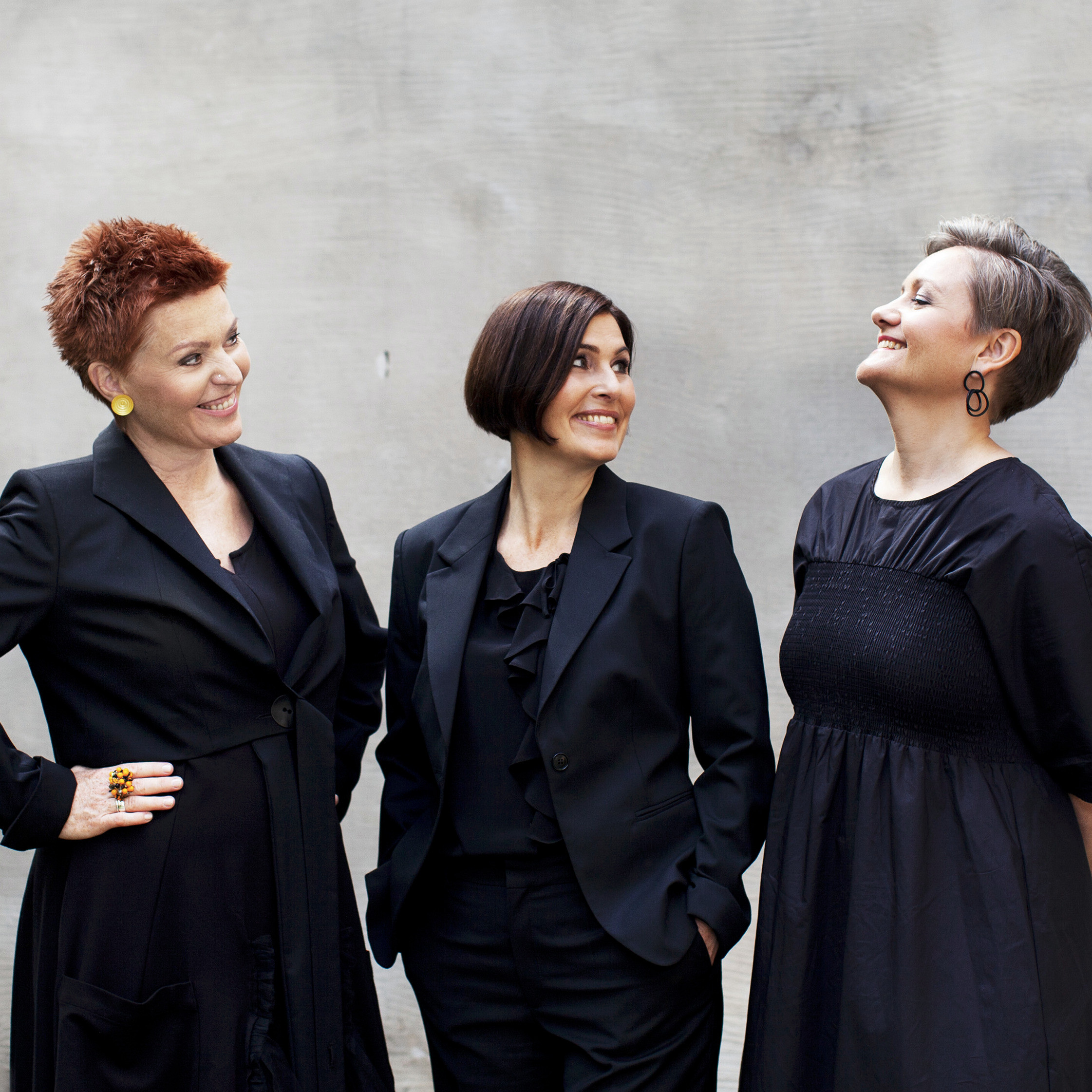 The three women of Trio Mediaeval dressed in black clothes looking at each other smiling.