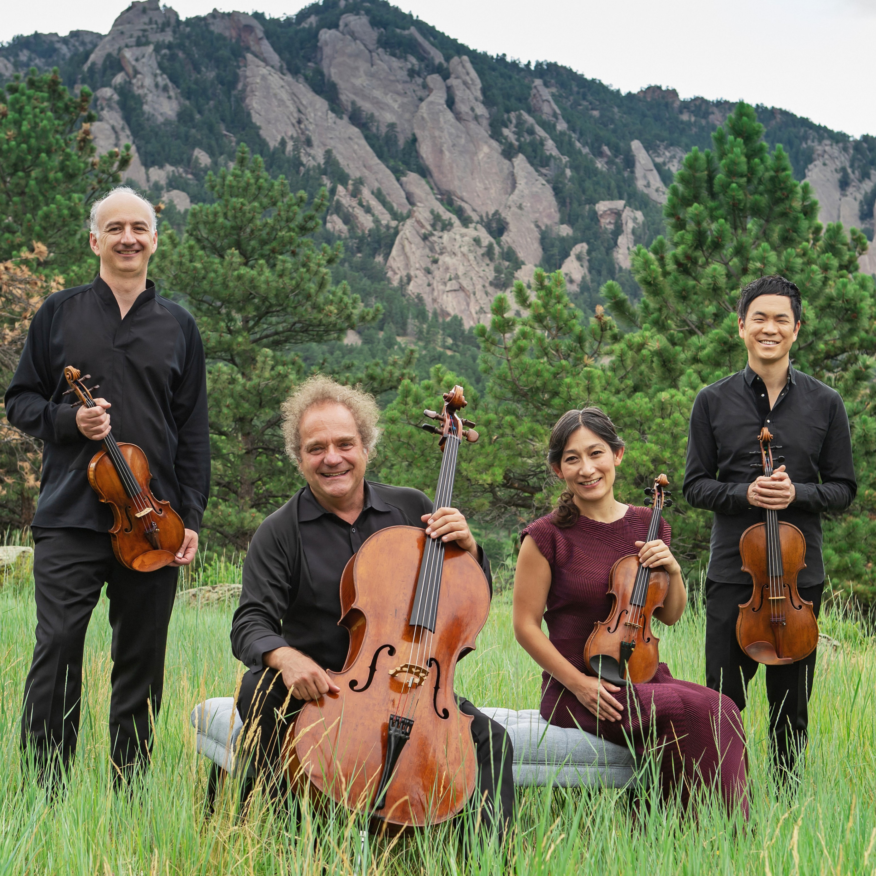 All four members of the quartet posing with their instruments outside in a grassy field with pine trees and a rocky boulder/mountian behind them.