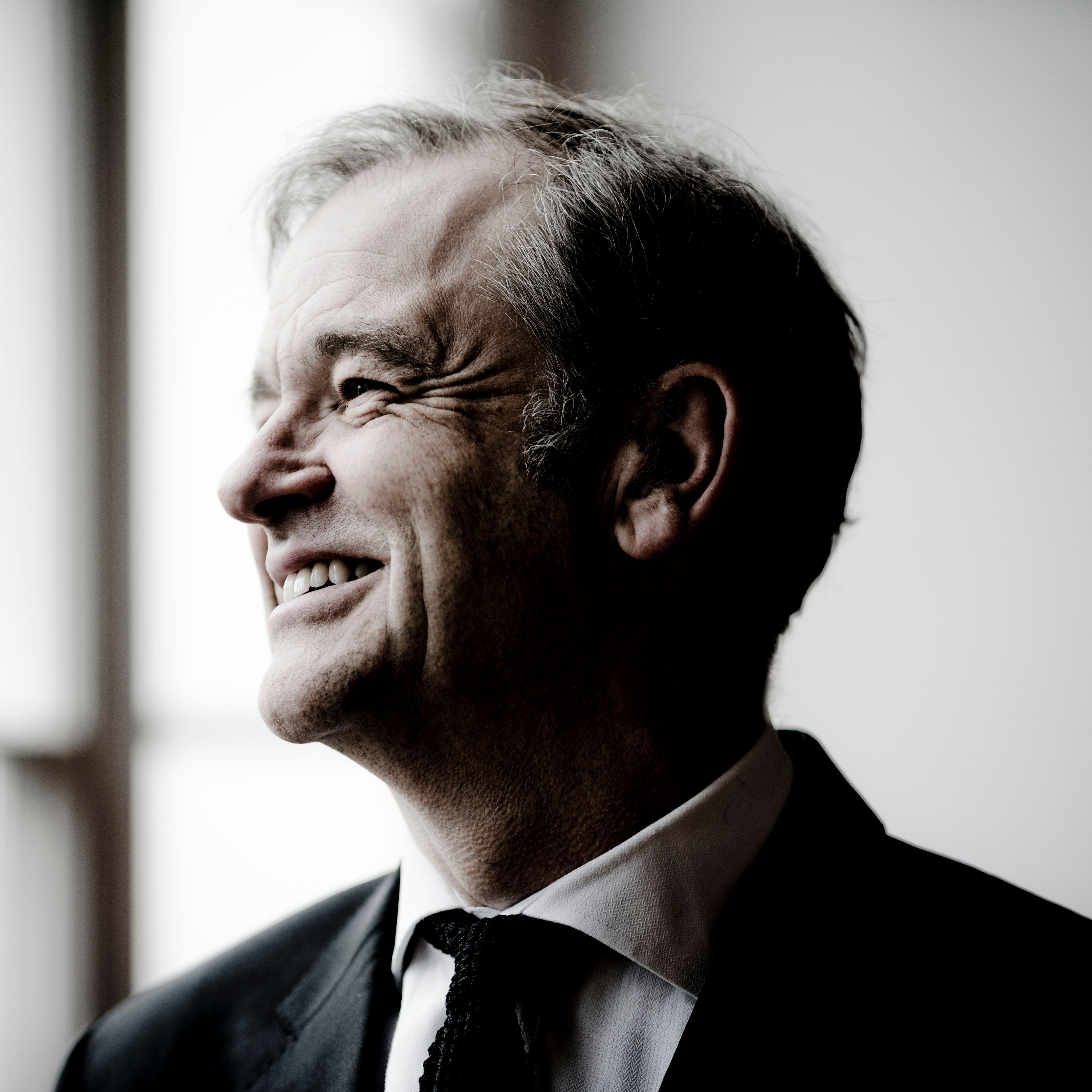 Closeup of Julius Drake dressed in a black blazer and tie with a white shirt, smiling, looking away to the left.