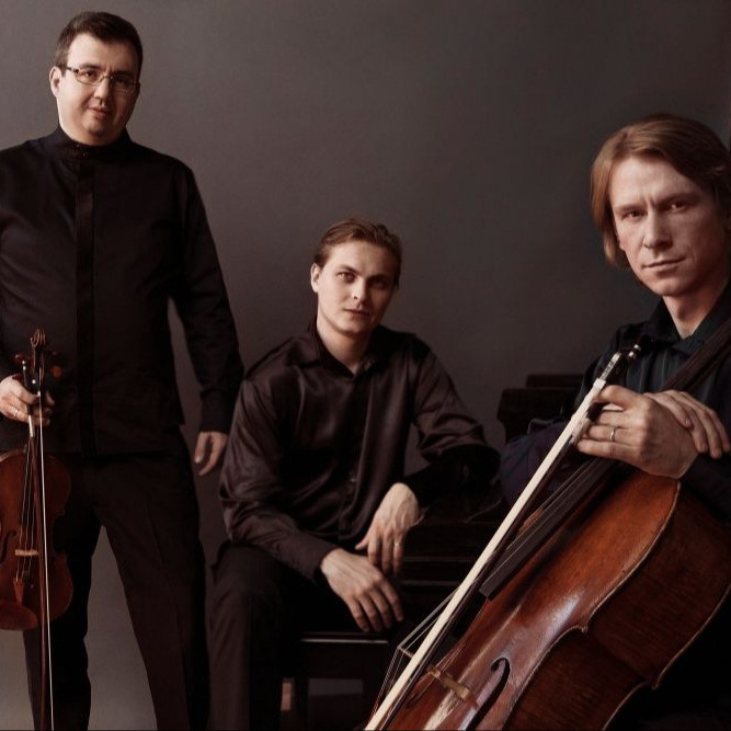 Members of the Hermitage Piano Trio posing with their instruments (cello, violin, and piano) in front of a dark gray background.