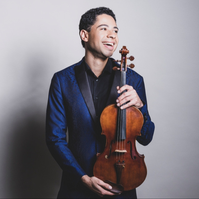 Jordan Bak wearing a black shirt and a dark blue blazer, smiling and holding his viola in front of a light gray background.