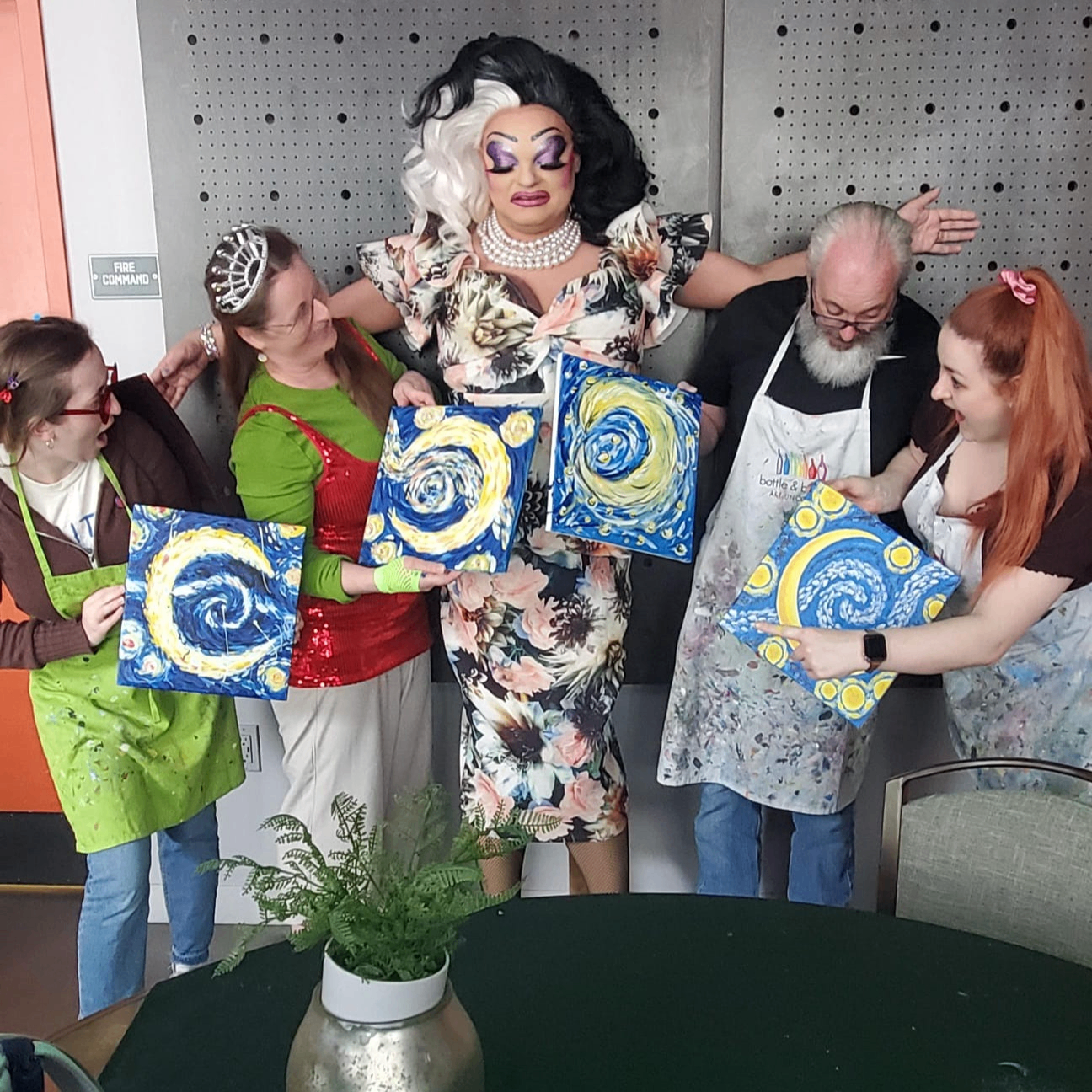 A drag queen frowns as she opens her arms to a group of people dressed in aprons, holding paintings of a Van Gogh inspired crescent moon. The party goers are smiling and making surprised faces.