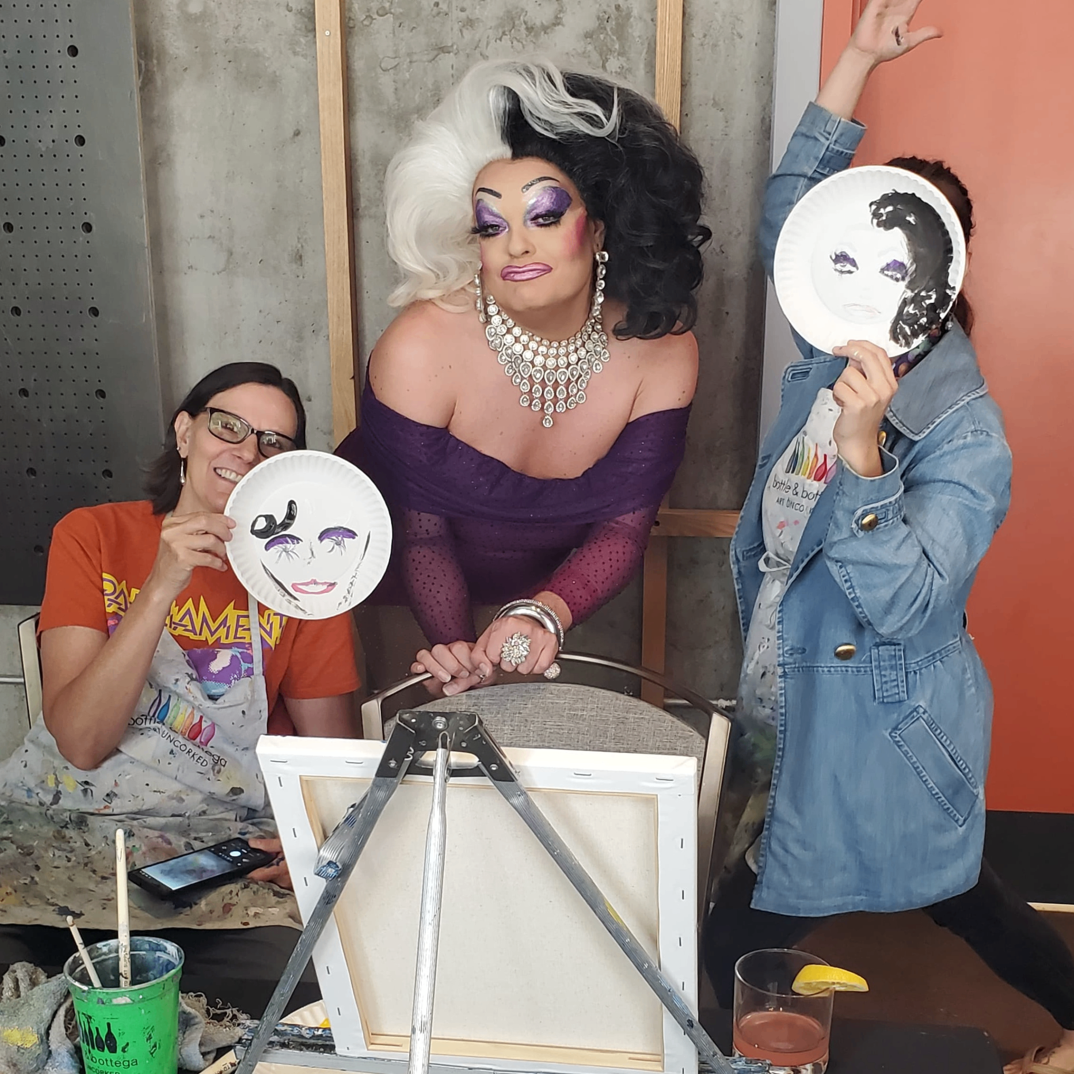 A drag queen stands in between two friends who painted the Drag queens face on paper plates. 