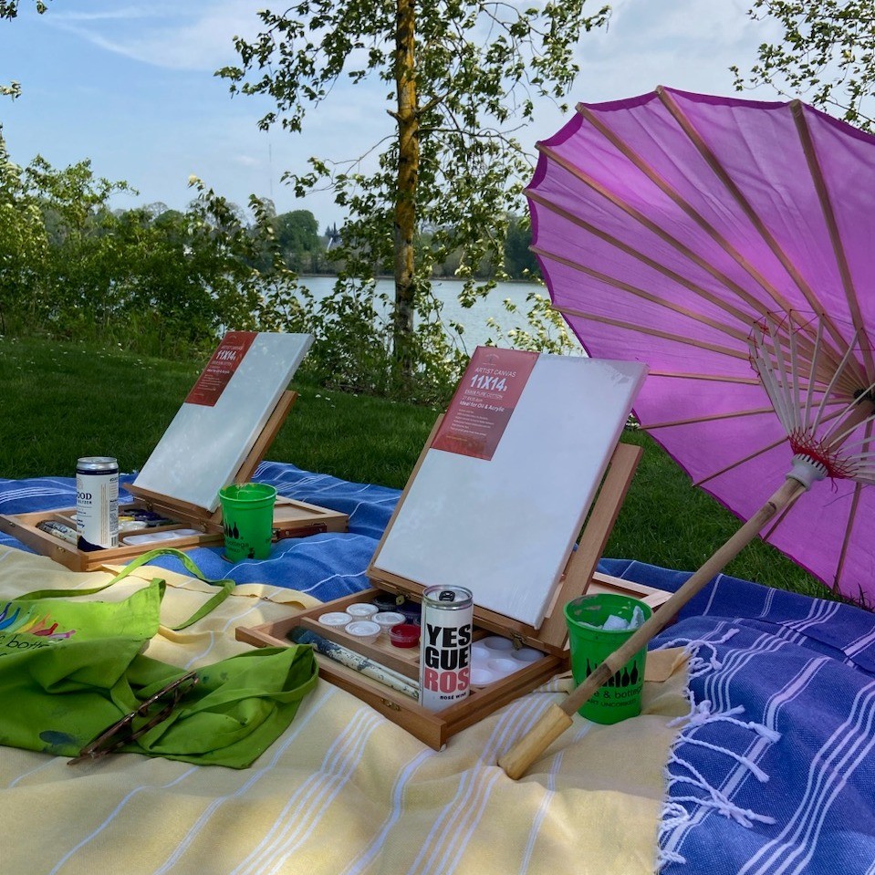 Two wooden easels with blank canvases, cans of wine, a pink parasol, and a green apron sit on colorful blankets in front of a grass lined river