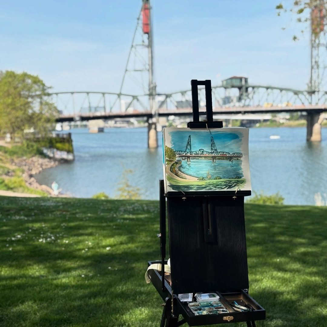 an easel set up in front of a river with a painting of the bridge in the distance