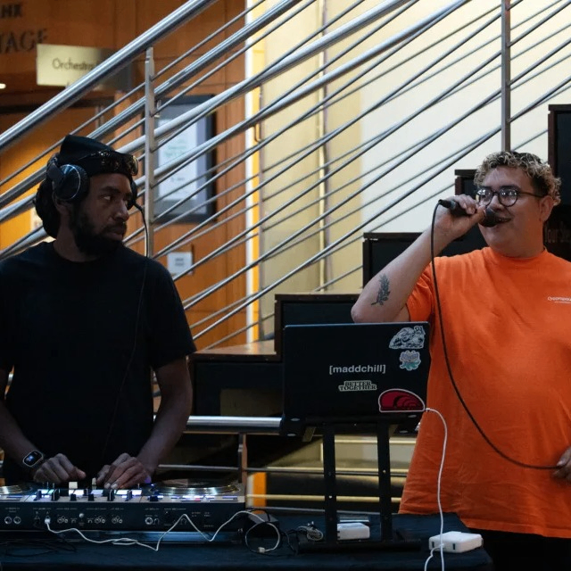 person speaking into a microphone standing next to a DJ and turntables