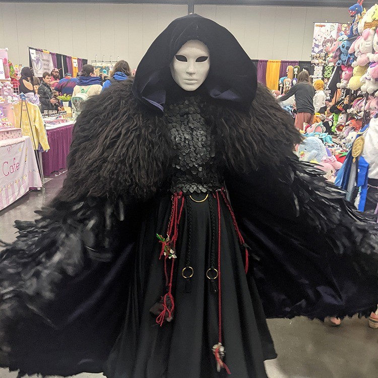 person in cosplay with a white mask and black feathered cloak